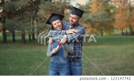 A joyful couple, both wearing graduation caps, shares a loving embrace while surrounded by a beautiful scenic park 123147742