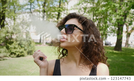 In a vibrant park, a stylish woman smokes leisurely, radiating a sense of confidence and relaxation effortlessly 123147743