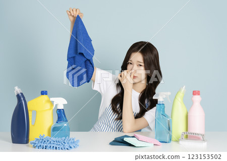 A woman in her 20s sitting in front of a table with cleaning tools, holding a mop and covering her nose 123153502