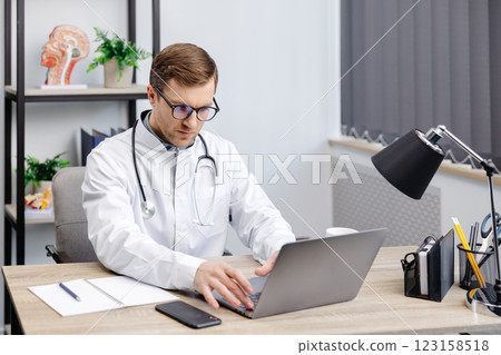 Doctor man sitting at the desk at his working place and working on laptop. Perfect medical service in clinic. Happy future of medicine and healthcare Doctor man sitting at the desk at his working place and working on laptop. Perfect medical service in clinic. Happy future of medicine and healthcare 123158518