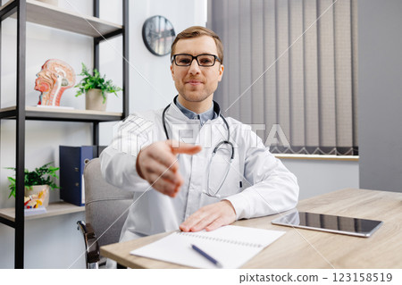 A young doctor in glasses and a white uniform sits at a desk and extends his hand to greet a patient A young doctor in glasses and a white uniform sits at a desk and extends his hand to greet a patient 123158519
