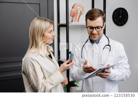 Male doctor holding in hand an aerosol inhaler on a demonstration to a female patient at his office Male doctor holding in hand an aerosol inhaler on a demonstration to a female patient at his office 123158530