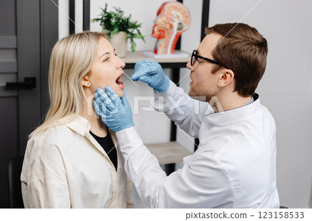 Doctor using inspection spatula to examine patient throat. ENT doctor doing throat exam of a woman. Patient opened her mouth to throat check-up Doctor using inspection spatula to examine patient throat. ENT doctor doing throat exam of a woman. Patient opened her mouth to throat check-up 123158533
