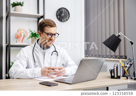 Portrait of man doctor talking to online patient on laptop screen sitting at clinic office desk giving online consultation for domestic health treatment. Telemedicine remote medical appointment Portrait of man doctor talking to online patient on laptop screen sitting at clinic office desk giving online consultation for domestic health treatment. Telemedicine remote medical appointment 123158559