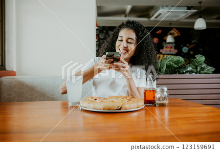 Afro girl taking a picture of a pizza with the phone in a pizzeria. Smiling young woman using phone photographing a pizza in a restaurant 123159691