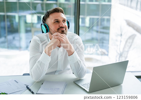 Thoughtful Professional Wearing Headphones Working at Desk in Modern Office Thoughtful Professional Wearing Headphones Working at Desk in Modern Office 123159806