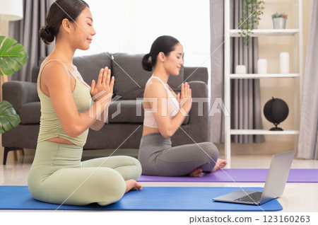 Two women doing yoga meditating exercise in living room, sitting in Easy seat pose with mudra in  bedroom. Working out wearing sportive top and leggings with laptop on blue mat. Relaxation and 123160263