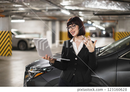 Caucasian businesswoman in indoor parking lot leaning on car with laptop conducting online video call, expressing ideas. Young woman in professional attire engaged in remote work. Caucasian businesswoman in indoor parking lot leaning on car with laptop conducting online video call, expressing ideas. Young woman in professional attire engaged in remote work. 123160628