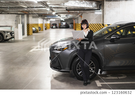 Caucasian female business professional stands in parking garage holding laptop. Young adult focused on work. Business attire, urban setting, technology, and modern lifestyle depicted. Caucasian female business professional stands in parking garage holding laptop. Young adult focused on work. Business attire, urban setting, technology, and modern lifestyle depicted. 123160632