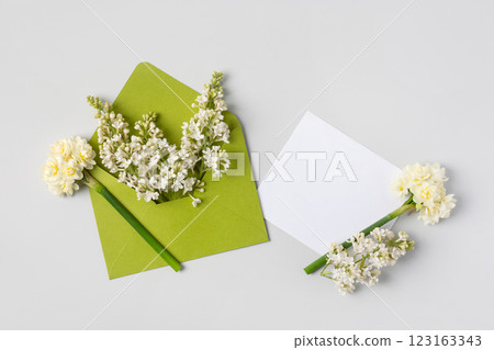 Green envelope with clean white sheet of paper for your text and spring flowers white lilac and white daffodils on gray background. Invitation to the holidays. Flat lay. 123163343
