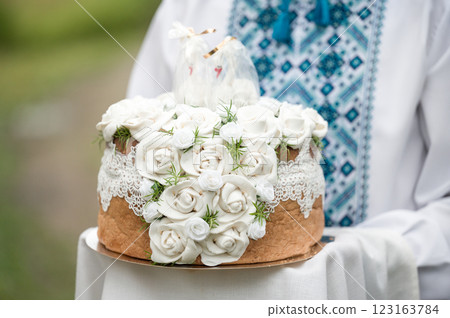 A beautifully decorated loaf of bread, adorned with white roses and lace, is held by a person wearing a traditional shirt. A beautifully decorated loaf of bread, adorned with white roses and lace, is held by a person wearing a traditional shirt. 123163784