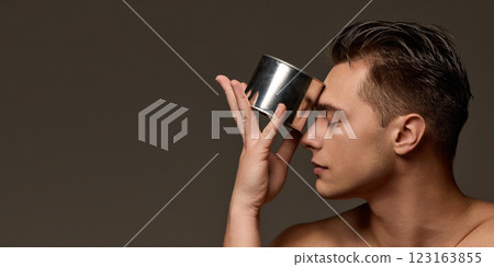 Young man pressing metallic container to forehead, eyes closed, illustrating self-care and deep skincare routine, against brown studio background. 123163855