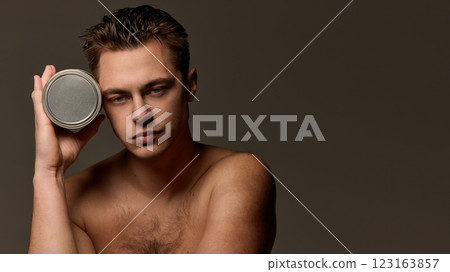 Portrait of young attractive, well-groomed man holding metallic container for skin-care product near head against brown studio background. 123163857