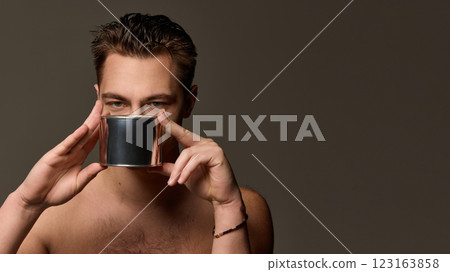 Shirtless man holding metallic container in both hands, looking through it with focused gaze against brown studio background. Shirtless man holding metallic container in both hands, looking through it with focused gaze against brown studio background. 123163858