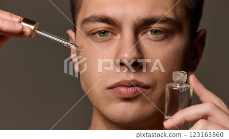 Close-up of man applying serum under eye with dropper, liquid running down cheek, focused expression against brown studio background. 123163860
