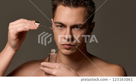 Shirtless man with serious expression applying serum with dropper near eye, looking directly at camera against brown studio background. 123163861