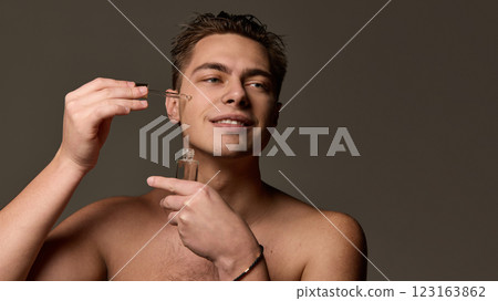 Man applying serum with dropper to cheek while holding glass bottle, smiling softly, looking to distance against brown studio background. 123163862