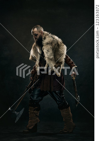 Viking warrior standing in battle stance gripping axe and sword, wearing fur cloak and leather armor against dark background. 123163872