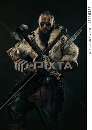 Fierce Viking warrior with war paint gripping two axes, dressed in fur cloak and chainmail, staring intensely against dark background. 123163874