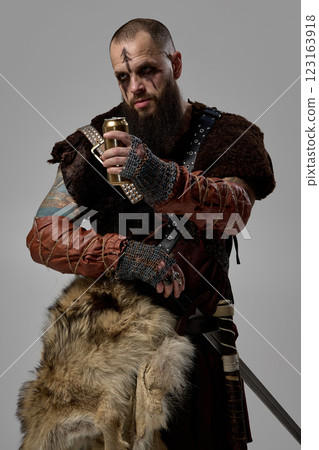 Portrait of Viking warrior gripping golden beer bottle, wearing fur cloak and battle-worn leather armor against studio background. 123163918