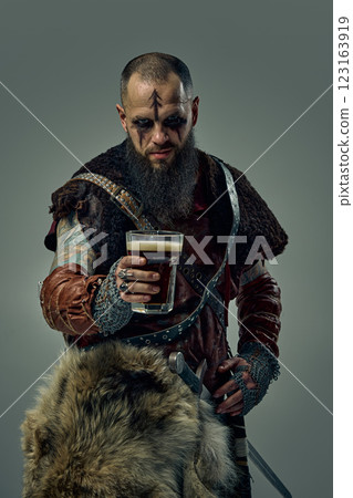Portrait of medieval Viking warrior dressed in fur cloak and leather armor, standing with sword, holding glass of beer, against studio background. 123163919