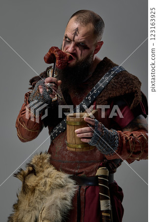 Mas as medieval warrior, Viking with intense gaze biting raw meat while gripping wooden mug filled with beer against studio background. Mas as medieval warrior, Viking with intense gaze biting raw meat while gripping wooden mug filled with beer against studio background. 123163925