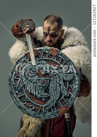 Man dressed as Viking warrior in medieval fur cloak, gripping sword while holding decorated shield with Norse engravings against studio background. Man dressed as Viking warrior in medieval fur cloak, gripping sword while holding decorated shield with Norse engravings against studio background. 123163927