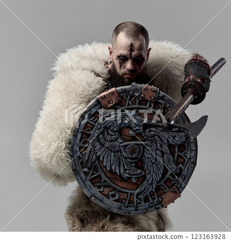 Viking warrior with decorated round shield with Norse symbols, battle axe raised, wearing fur cloak and leather armor against studio background. Viking warrior with decorated round shield with Norse symbols, battle axe raised, wearing fur cloak and leather armor against studio background. 123163928