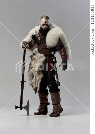 Man dressed as Viking warrior in medieval fur cloak and rugged armor, resting heavy axe on ground while looking down in contemplation 123163932