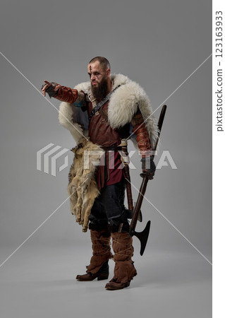Portrait of man as medieval Viking warrior dressed in fur cloak and leather armor, standing with axe and pointing forward against studio background 123163933