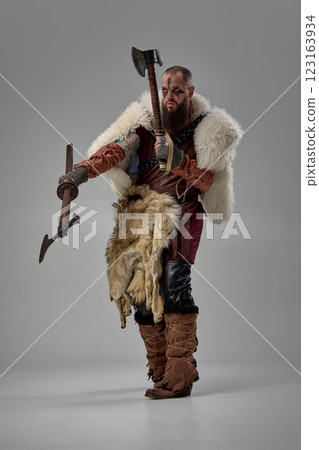 Warrior gripping two axes with intense gaze, man dressed in armor and fur cloak enhancing battle-ready stance against grey studio background. 123163934