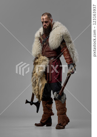Full-length portrait of fierce man, medieval warrior, Viking in full armor and fur elements, standing with two axes against grey studio background Full-length portrait of fierce man, medieval warrior, Viking in full armor and fur elements, standing with two axes against grey studio background 123163937