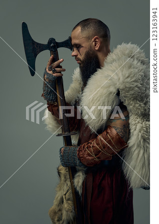 Portrait of man dressed as medieval warrior, Viking standing with eyes closed, with head near the age, against grey studio background. Portrait of man dressed as medieval warrior, Viking standing with eyes closed, with head near the age, against grey studio background. 123163941