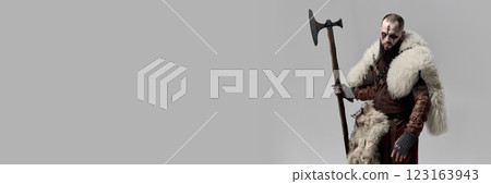 Portrait of medieval man, Viking in leather armor and fur cloak, standing with axe and gazing with determination against grey studio background. Portrait of medieval man, Viking in leather armor and fur cloak, standing with axe and gazing with determination against grey studio background. 123163943