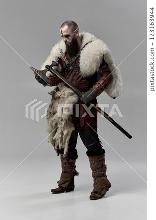 Full-length portrait of serious man, medieval Viking in leather armor and fur cloak, holding his axe against grey studio background. Full-length portrait of serious man, medieval Viking in leather armor and fur cloak, holding his axe against grey studio background. 123163944