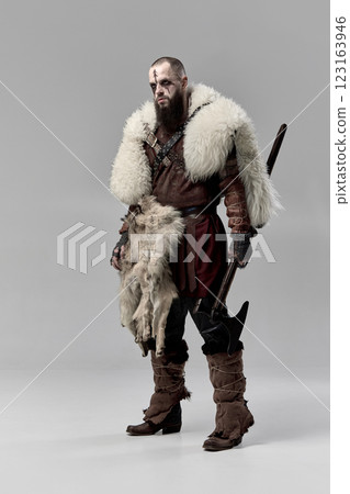 Full-length portrait of determined man, medieval Viking dressed in leather armor and fur cloak, standing with axe against grey studio background 123163946