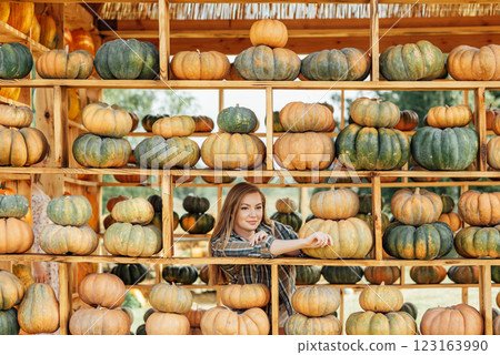 young woman stands at pumpkin festival, holding large orange and green pumpkin. She is surrounded by rows of pumpkins and a wooden display. The autumn farm setting captures the harvest season vibe. young woman stands at pumpkin festival, holding large orange and green pumpkin. She is surrounded by rows of pumpkins and a wooden display. The autumn farm setting captures the harvest season vibe. 123163990