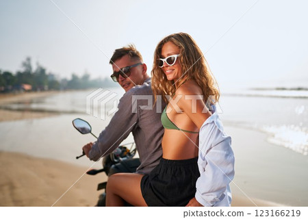 Smiling couple on motorbike at sunny beach. Young man and woman relax on motorcycle near ocean. Romantic adventure, love, travel by scooter, enjoy summer vibes, palm trees in background. Smiling couple on motorbike at sunny beach. Young man and woman relax on motorcycle near ocean. Romantic adventure, love, travel by scooter, enjoy summer vibes, palm trees in background. 123166219