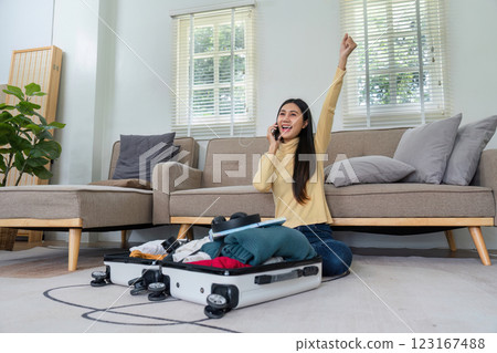 Cheerful Young Woman Packing Suitcase for Travel While Talking on Phone in Modern Living Room 123167488