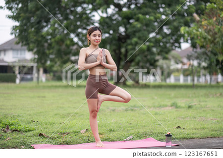 Young Woman Practicing Yoga in the Park on a Sunny Day, Standing in Tree Pose on a Pink Mat with Greenery in the Background 123167651