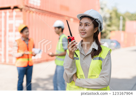 Focused female construction supervisor using a walkie-talkie while her team collaborates at a shipping container site 123167904