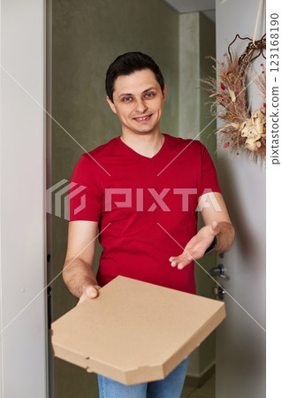 positive delivery man in red t-shirt giving food order, holding pizza box standing at doorway positive delivery man in red t-shirt giving food order, holding pizza box standing at doorway 123168190