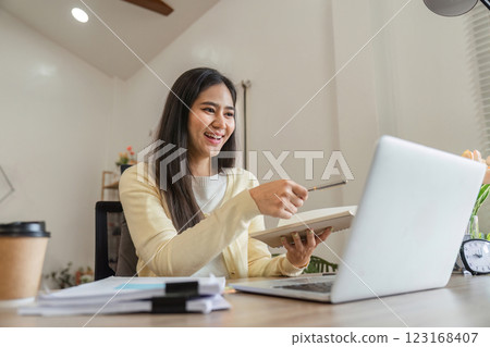 Cheerful Asian woman presenting notes while working on a laptop, highlighting engagement and creativity in her home office. 123168407