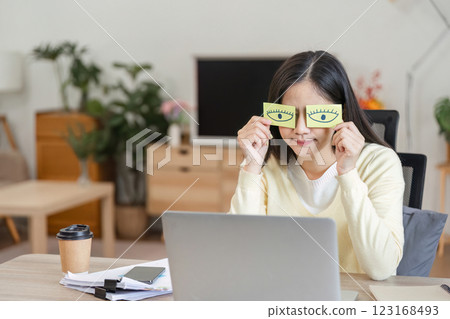 Playful Asian woman holding paper eyes while working on a laptop, showcasing creativity and fun in the home office. Playful Asian woman holding paper eyes while working on a laptop, showcasing creativity and fun in the home office. 123168493