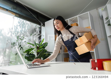E-commerce businesswoman managing orders on a laptop while carrying packages in a modern office. E-commerce businesswoman managing orders on a laptop while carrying packages in a modern office. 123168494
