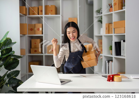 Excited young Asian woman celebrating success in e-commerce while holding packages in her workspace. Excited young Asian woman celebrating success in e-commerce while holding packages in her workspace. 123168618