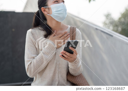 Young woman wearing a mask, holding smartphone, and showing emotion outdoors. Young woman wearing a mask, holding smartphone, and showing emotion outdoors. 123168687