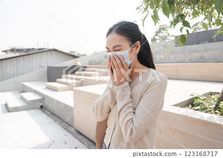 Young woman wearing a mask covering her face with hands outdoors, expressing emotion. 123168717