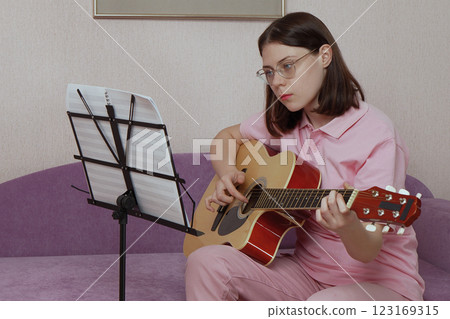 Young female guitarist playing and learning acoustic guitar sitting at sofa in home interior 123169315