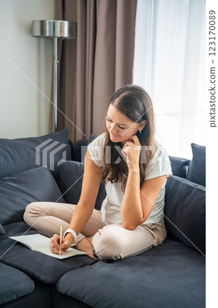 Young woman siting on the sofa and writing her personal diary, writing down thoughts, dreams and plans as part of psychotherapy after working with neurocoaching Young woman siting on the sofa and writing her personal diary, writing down thoughts, dreams and plans as part of psychotherapy after working with neurocoaching 123170089